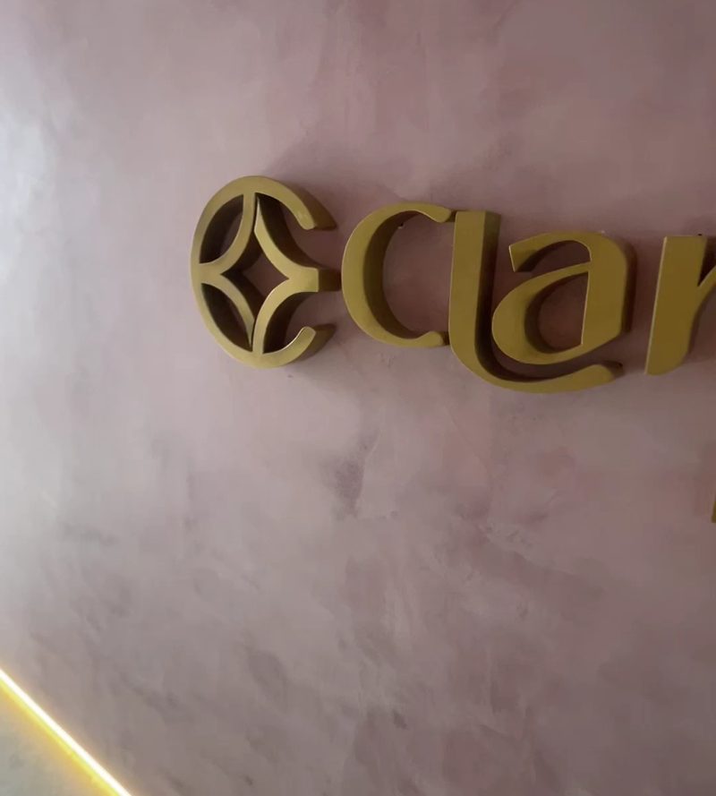 Letreiro dourado da Clara Vida na parede rosada, com fita de luz